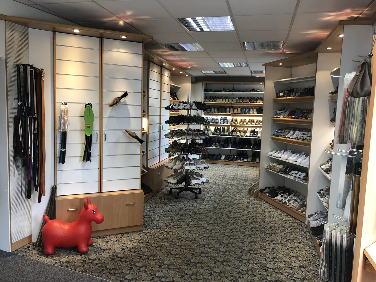 • Abgebildet ist ein Ladengeschäft mit Regalen, in den Auslagen befinden sich unterschiedliche Schuhe sowie diverse Einrichtungselemente und ein rotes Kinderpferd ist zu sehen.